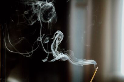 A close-up of smoke rising from an incense stick, creating a swirling pattern against a dark background.