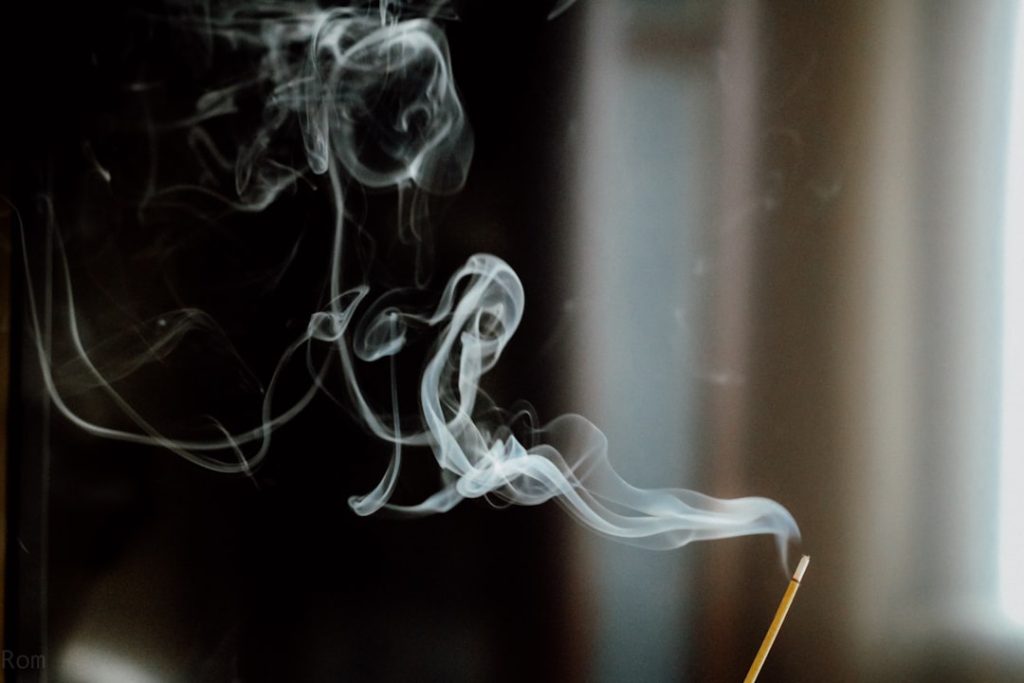 A close-up of smoke rising from an incense stick, creating a swirling pattern against a dark background.