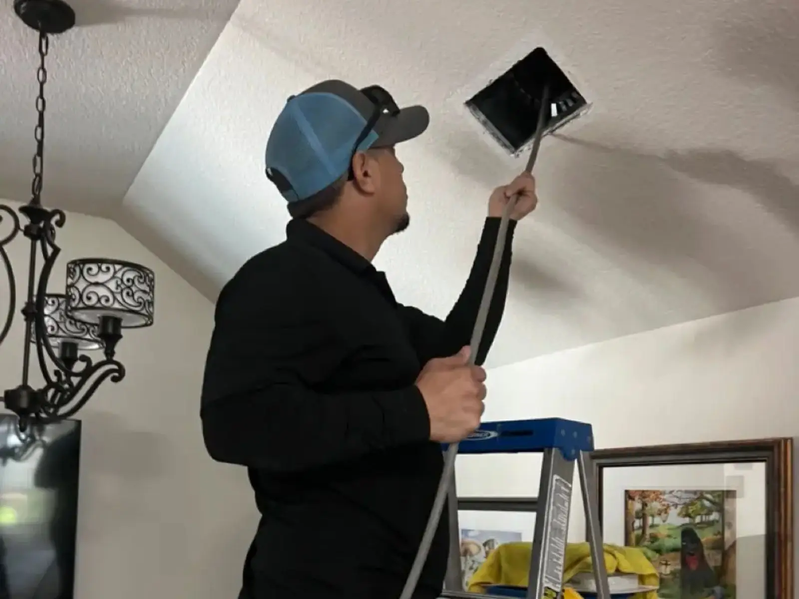 A man on a ladder cleaning the ceiling with a cloth, focused on removing dust and cobwebs.