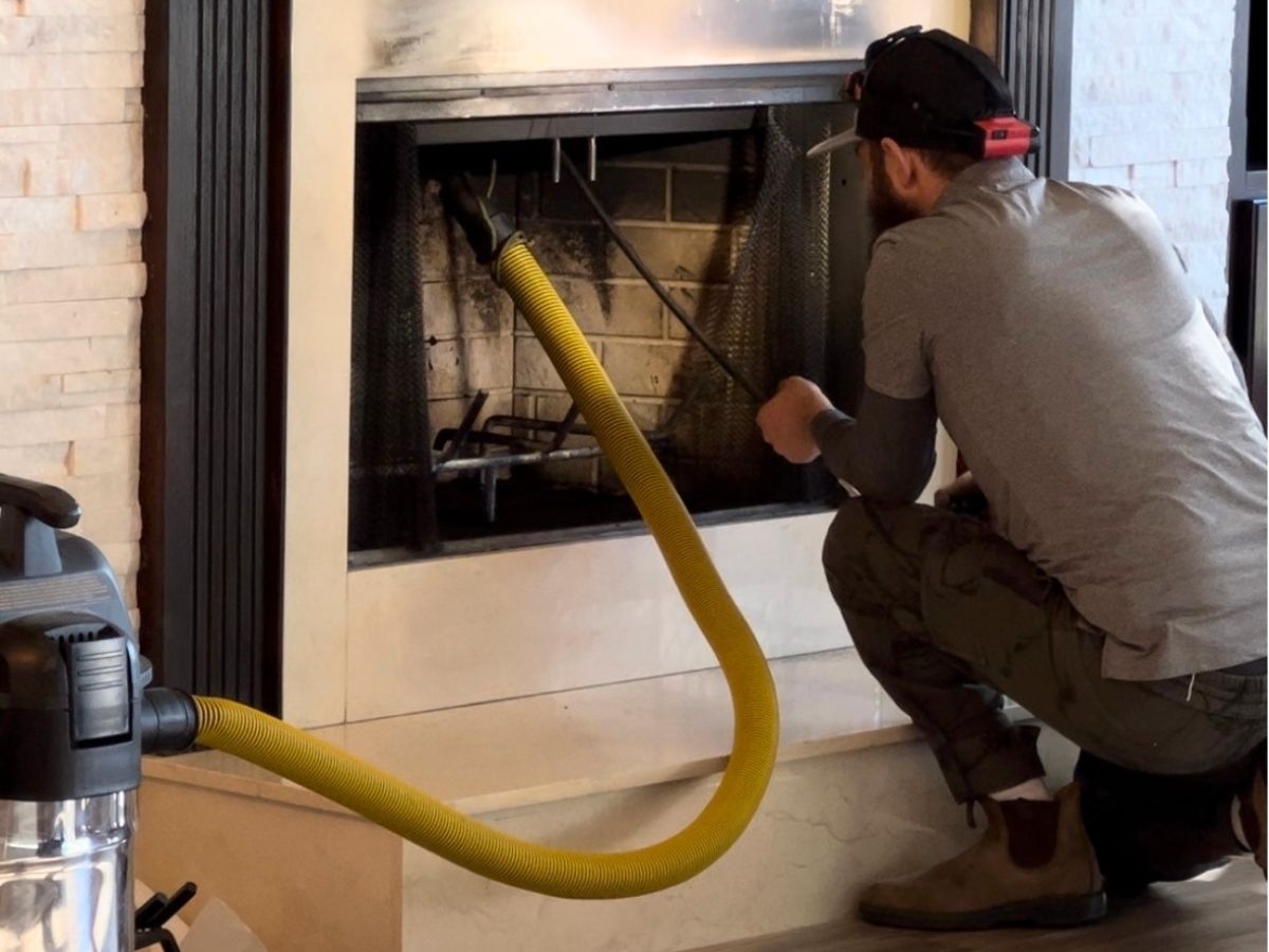 Technician performing a professional chimney sweep service, cleaning soot and debris from a residential fireplace to improve safety and indoor air quality, with vacuum equipment and protective gear visible.
