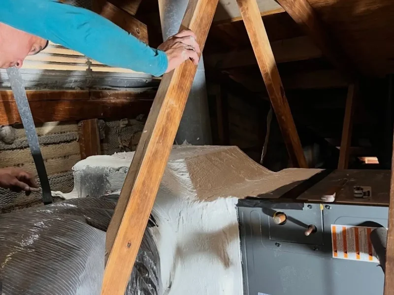Technician applying white mastic sealant to the duct connections of an HVAC air handler in a residential attic.