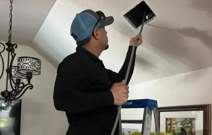 A man on a ladder cleaning the ceiling with a cloth, focused on removing dust and cobwebs.