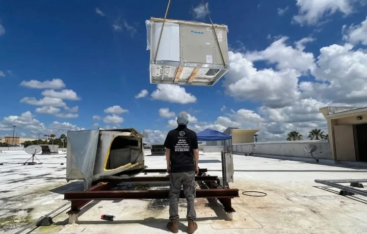 A large Daikin commercial HVAC unit suspended in mid-air by a crane above a rooftop.