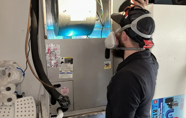 HVAC technician wearing a respirator and headlamp inspecting a blower motor illuminated by UV light.