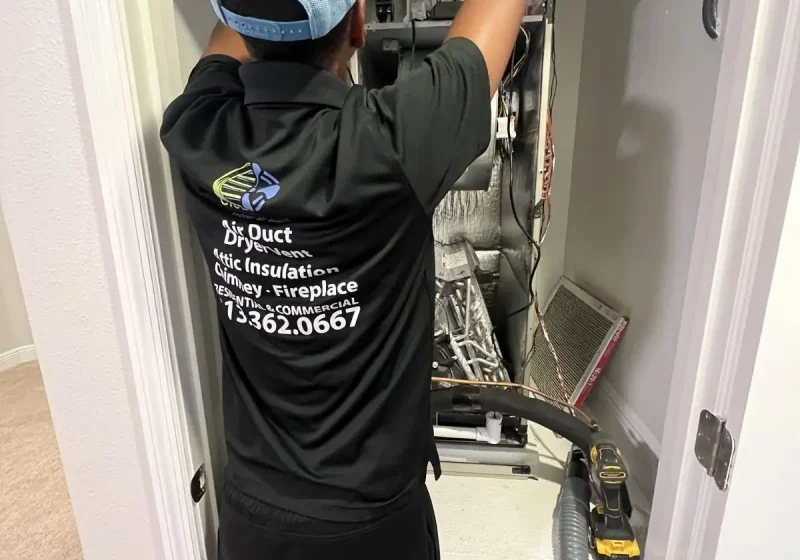 Rear view of an HVAC technician in a black company shirt working on an indoor air handler inside a utility closet.